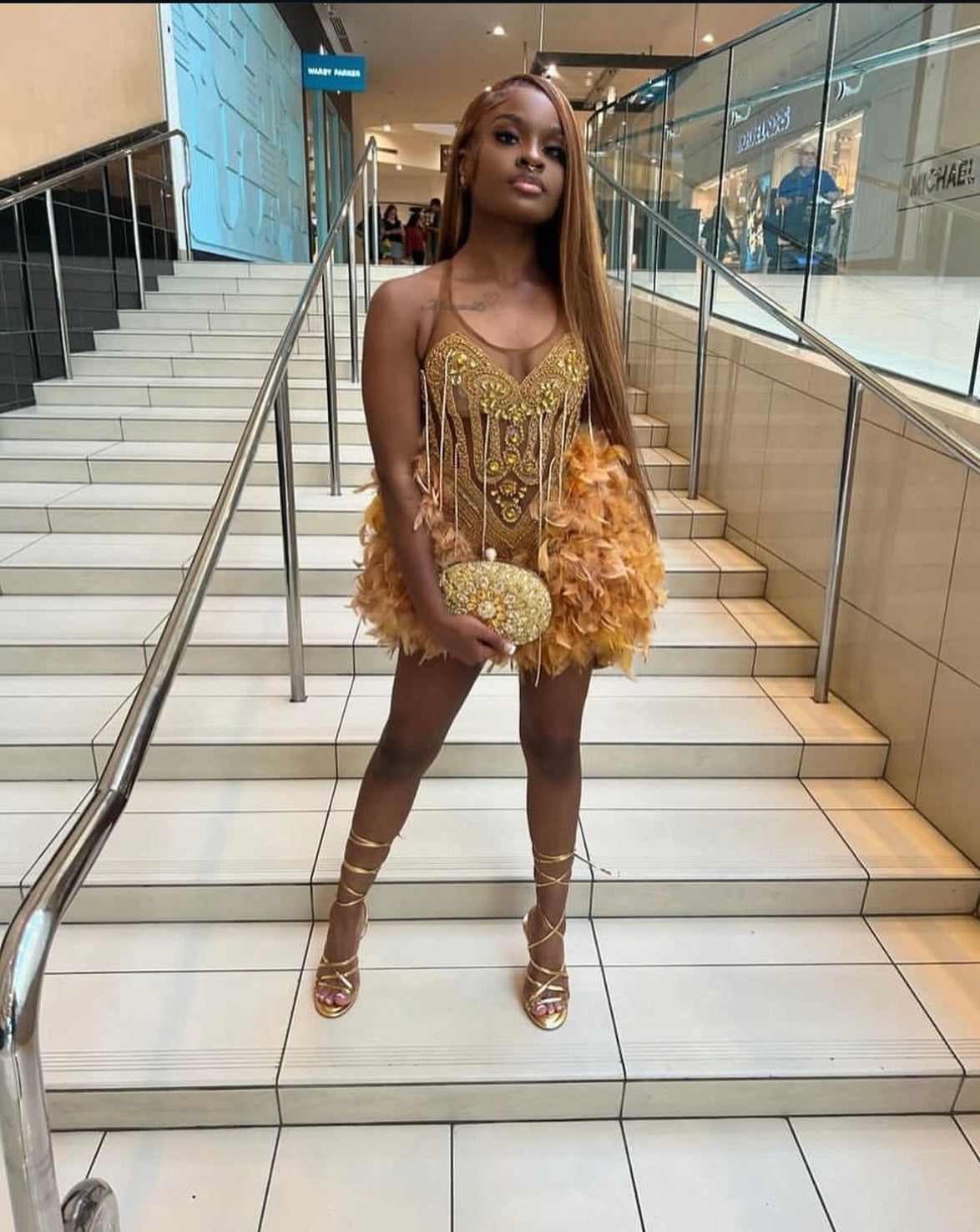 Black woman with light brown long hair and a gold dress with feather trim. Posing on mall steps. 